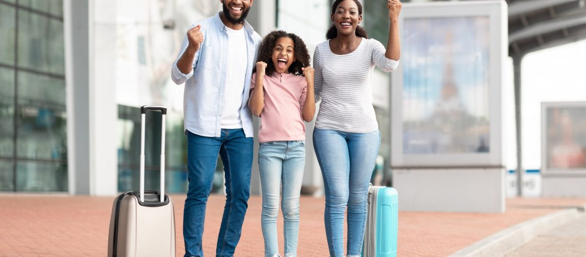 Summer Vacation Concept. Cheerful positive black family of three people having fun and shaking clenched fists, making winner gesture, travelling together standing near airport terminal with baggage