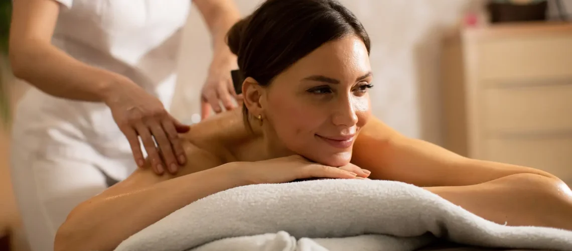 A woman lies on a massage table while a therapist applies pressure to her back. The scene shows a spa interior with calming colors. It is daytime, and the light is soft.